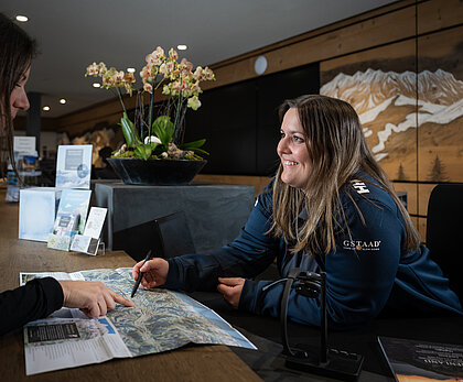 <p>Eine Mitarbeiterin von Gstaad Saanenland Tourismus zeigt einem weiblichen Gast etwas auf einer Panoramakarte. Im Hintergrund die Schalter des Tourismusbüros Gstaad.</p>