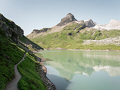 Der Sanetschsee scheint in einer grünen Farbe und die Berge spiegeln sich darin. Auf der linken Seite sieht man den schmalen Wanderweg am steilen Ufer entlang. Diese Seite liegt im Schatten während auf dem Rest des Bildes die Sonne scheint und der Himmel hellblau ist.