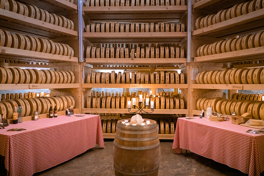 <p>Hintergrund mit Regalen voller Käse, in der Mitte stehen Tische bereit für die Degustation.</p>