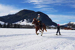 Person reitet auf einem Pferd über ein verschneites Feld, daneben eine weitere Person auf Skiern; Berge und Höfe im Hintergrund.
