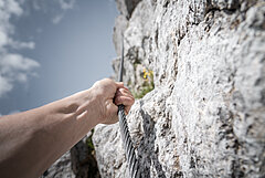 Eine Hand hält ein Kletterseil an einer Felswand, blauer Himmel im Hintergrund.