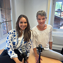 Zwei lachende Frauen mit Podcast Mikrofon sitzen auf dem Tisch und lachen in die Kamera.