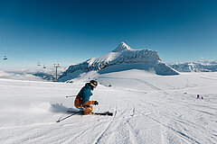 A skier in an orange and turquoise ski suit going down a ski slope beside a chairlift, with a view of the glacier and the mountain panorama.
