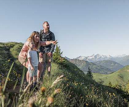 Eine Familie steht auf einem Wanderweg in alpiner Landschaft und blickt auf die umliegenden Berge. Grüne Wiesen und ein weiter Ausblick sind im Hintergrund zu sehen.