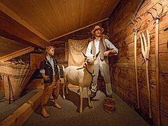A white Saanen goat and two herdsmen stand together. On the right-hand side, pitchforks hang on the wall