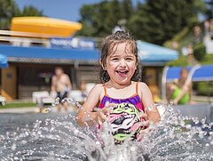 Ein kleines lachendes Mädchen in buntem Badeanzug spritzt in einem Freibad Wasser auf. Im Hintergrund Badegäste und ein Gebäude mit Terrasse mit gelbem Sonnenschirm.
