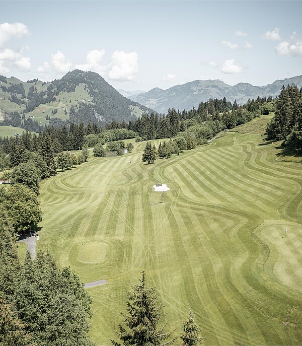 Eine malerische Golfanlage mit gepflegtem Rasen und sanften Hügeln. Im Hintergrund sind Berge und bewaldete Flächen sichtbar.