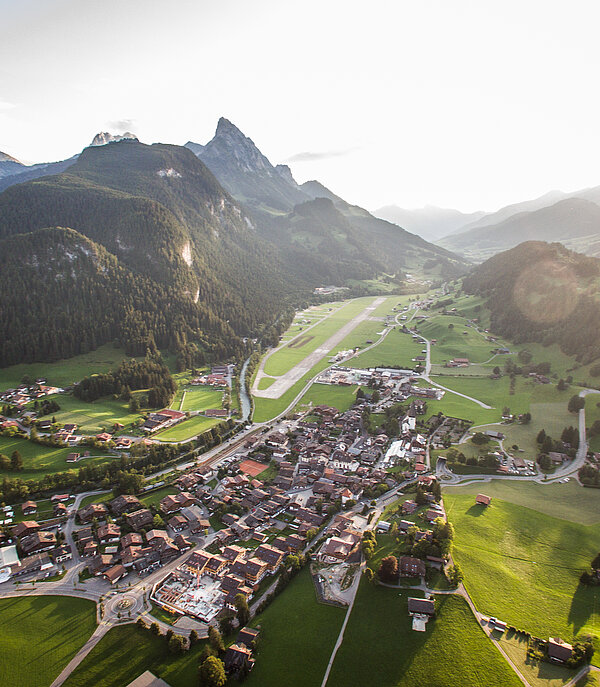 Sommer-Luftaufnahme vom Dorf Saanen und dem Flugplatz mit dem Le Rubli Gipfel und weiteren Bergen im Hintergrund.