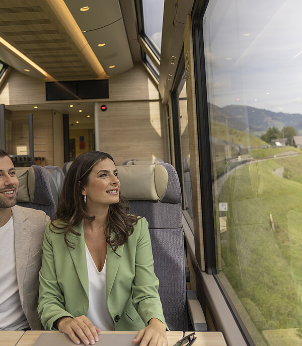 Anreise_mit_Zug.jpg Ein Mann und eine Frau sitzen in einem Zugabteil und schauen aus dem grossen Panoramafenster in die grüne Landschaft