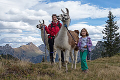 Zwei Lamas und eine Mutter mit ihrer Tochter stehen auf einem Berg und schauen in die Kamera.