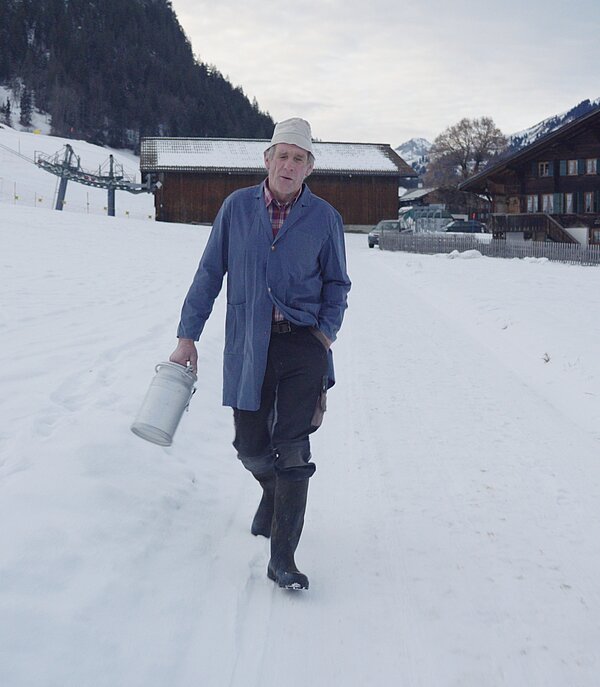 Ein Mann läuft mit einer Milchkanne in der Hand über eine schneebedeckten Weg. Er bewegt sich auf die Kamera zu.