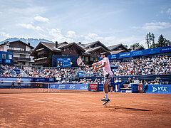 Dominic Thiem springt auf dem Tennisfeld und schlägt Ball zurück, im Hintergrund schaut das Publikum gespannt zu. Dahinter Häuser, Wälder und Berge, leicht bewölkter Himmel.
