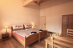 Wooden double bed with country-style bedlinen and two bedside tables with lamps. A table and two chairs stand on the right. In the background is a light wooden door and a dark brown wooden cupboard.
