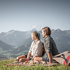 Eine Frau und ein Mann, welche auf einer Picknickdecke sitzen und eine Berglandschaft bewundern.
