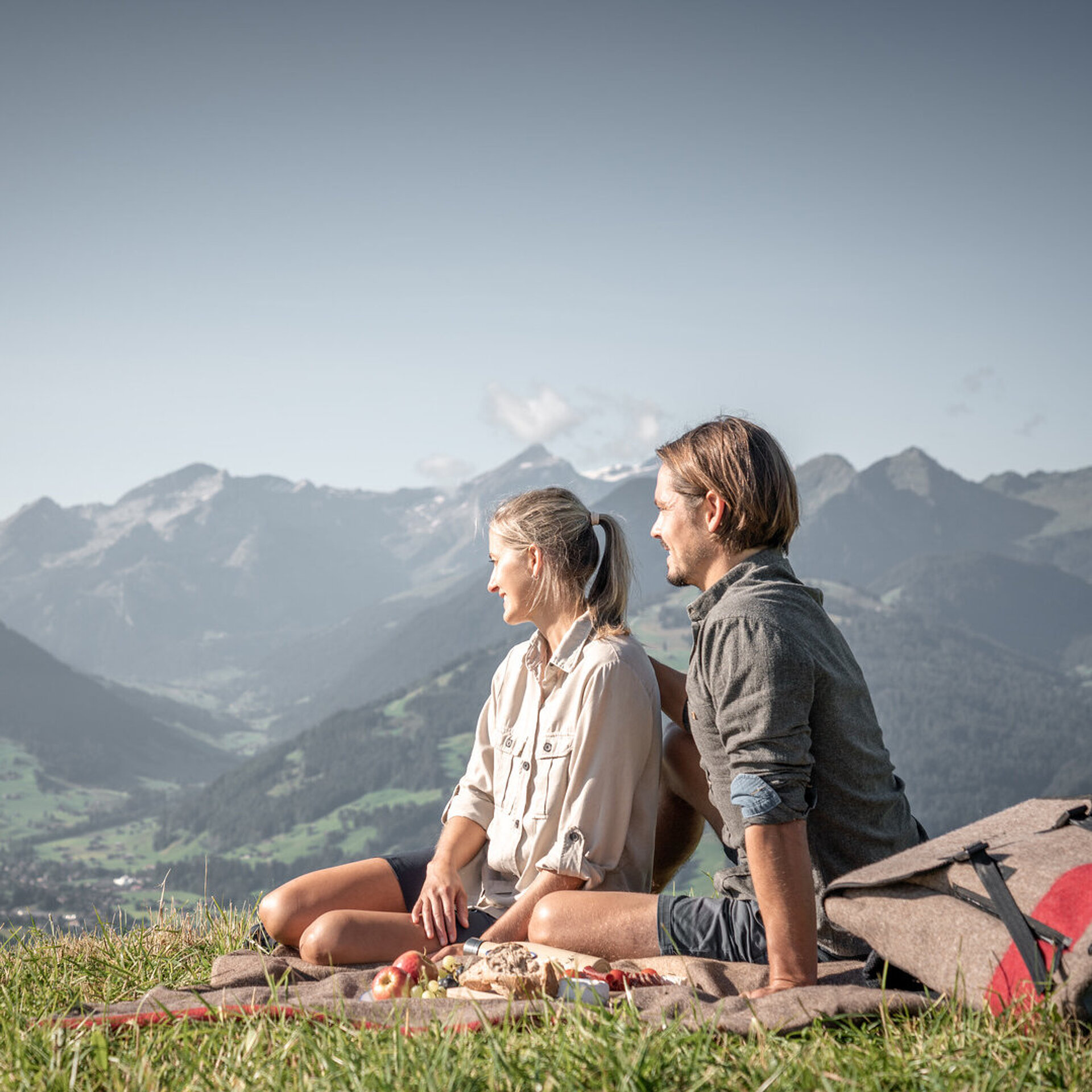 <p>Eine Frau und ein Mann, welche auf einer Picknickdecke sitzen und eine Berglandschaft bewundern.</p>