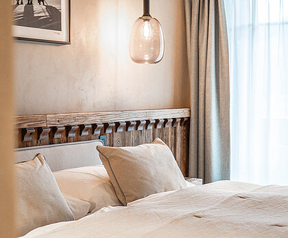 Zimmer im Hotel The Mansard Gstaad mit einem warmen beige und Grauton als Hauptfarbe. Das Zimmer ist beleuchtet und am Fenster ist ein transparenter Vorhang.