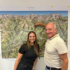 Zwei Personen stehen vor einer großen, detaillierten topografischen Karte von Gstaad. Die Person rechts zeigt mit ausgestrecktem Arm auf eine bestimmte Stelle auf der Karte, die verschiedene Berglandschaften und Täler darstellt.