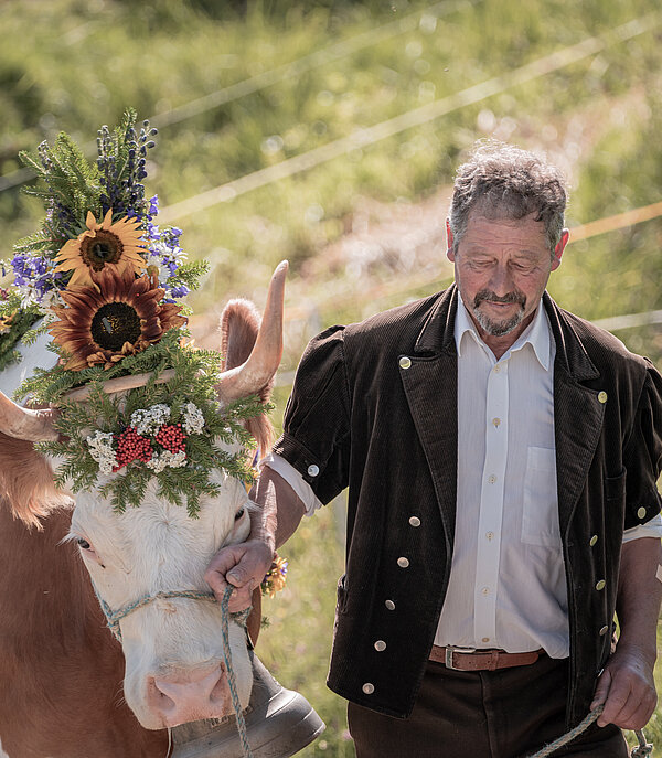 <p>Ein Bauer in traditioneller Alpkleidung mit schwarzer Jacke und weissem Hemd führt eine Kuh an der Hand. Die Kuh ist mit wunderschönen Blumen geschmückt.</p>