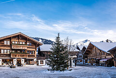 Dorfplatz in Saanen im Winter mit einer grünen Tanne in der Mitte. Rundum typische hölzerne Chalets.