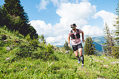 Un coureur de trail qui court vers l'appareil photo. Il évolue sur un sentier entre l'herbe et les pierres. À l'arrière-plan, on aperçoit un ciel bleu et de gros nuages blancs.
