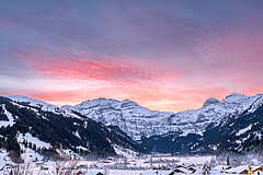 Winterliche Berglandschaft mit verschneiten Hängen und Dorf im Tal, beleuchtet von rosa Morgenhimmel und ersten Sonnenstrahlen.