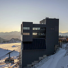 Dark, tall building. On the left, a cable car cabin and a chairlift. On the right, a wooden chalet and a bridge. Snow-covered mountains and the glacier in the background.
