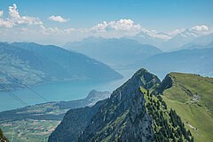 Summer view of Lake Thun with mountain peaks in the foreground and a panoramic mountain backdrop.