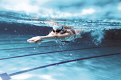 Une nageuse portant un bonnet de natation et des lunettes plonge dans une piscine couverte.