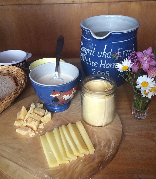 Auf einem Holztisch stehen ein Brotkorb, eine leere Tasse, eine Zuckerdose, ein Krug , eine Tasse mit Rahm, kleine Hobelkäsestückchen, Alpkäsestücke und in einem Glas gerollter Hobelkäse. Dazu steht ein kleiner Blumenstrauss in einem Glas.