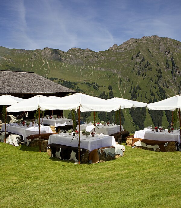 <p>Eine Gruppe von Tischen mit weissen Tüchern und Sonnenschirmen auf einer Wiese, eine Bergkette vor blauem Himmel.</p>