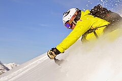 Person in yellow winter gear riding dynamically through powder snow on a snow bike, snow spraying up, blue sky and mountains in the background.