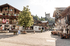 Paved road and square with benches and a tree. On the right a restaurant with tables and chairs outside. Several wooden chalets along the road. A castle-like hotel in the background.