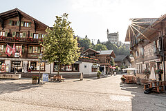 Gepflasterte Strasse und Platz mit Sitzbänken und einem Baum. Rechts ein Restaurant mit Tischen und Stühlen im Freien. Entlang der Strasse mehrere Holzchalets. Im Hintergrund ein schlossähnliches Hotel.