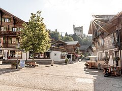 Rue pavée et place avec des bancs et un arbre. A droite, un restaurant avec des tables et des chaises en plein air. Le long de la route, plusieurs chalets en bois. En arrière-plan, un hôtel ressemblant à un château.