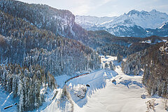 Un train circulant dans une vallée enneigée avec rivière, pentes boisées et paysage de montagne hivernal.