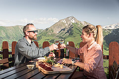 A couple sits on a sun terrace of a mountain restaurant, enjoys a cheese and meat platter and toasts with a glass of white wine, with a mountain panorama in the background.
