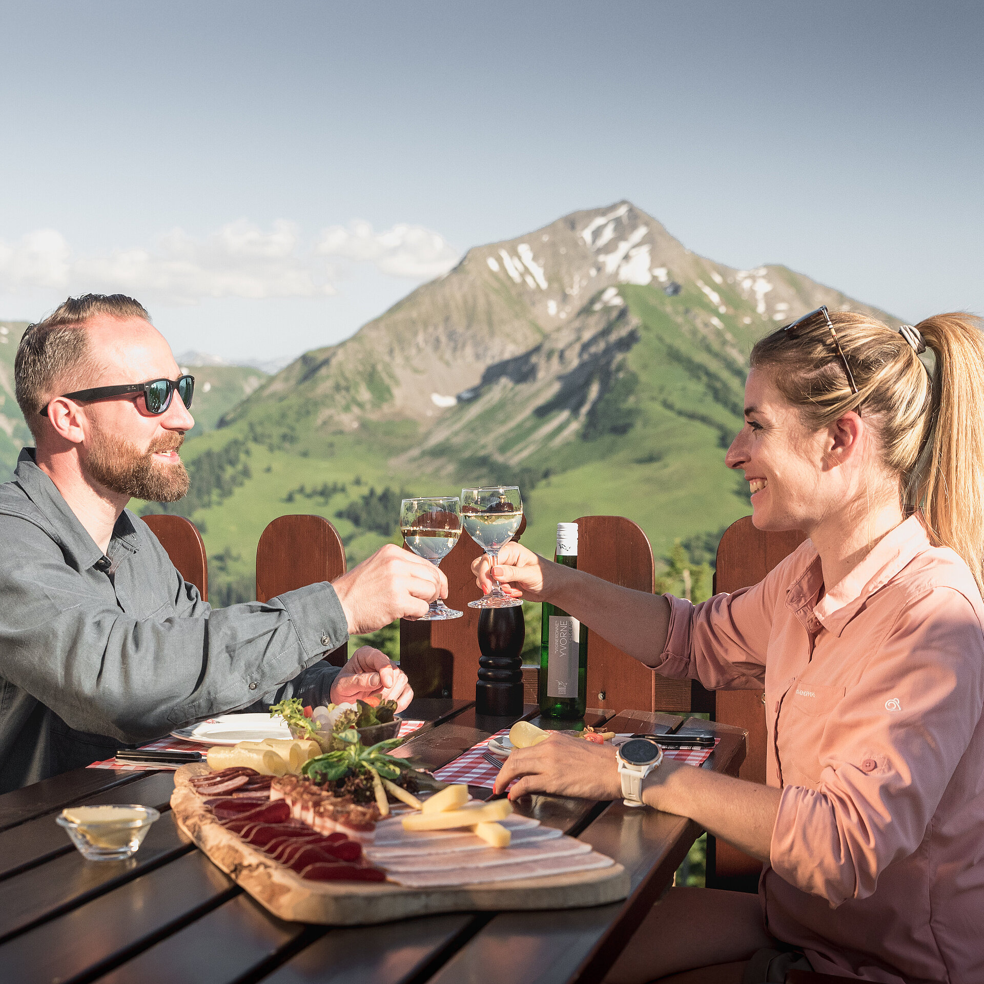 <p>Ein paar sitzt auf einer Sonnenterrasse eines Bergrestaurants, geniesst eine Käse- und Fleischplatte und stösst mit einem Glas Weisswein an. Im Hintergrund ein Bergpanorama.</p>