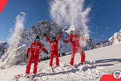 Drei Personen in roten Skijacken feiern im Schnee und werfen Schnee in die Luft. Im Hintergrund sind beeindruckende Berge und blauer Himmel zu sehen.