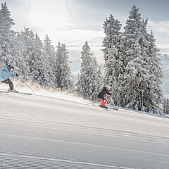 Two skiers on a freshly grooved slope with a view of snowy landscape.