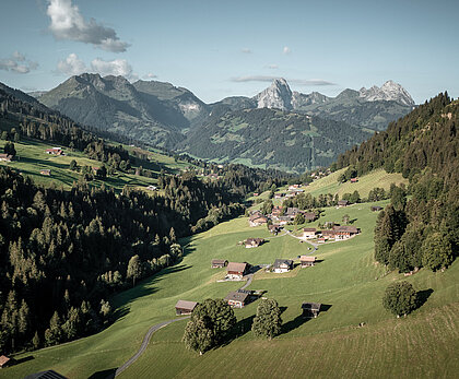 Der Blick ist etwas erhöht aus dem Turbachtal Richtung Gstaad - talauswärts. Im Tal sind Chalets und kleine Scheunen verteilt, rechts und links sind die Bergflanken mit Wald bedeckt. Zuhinterst sind felsige Gipfel zu sehen.