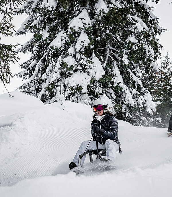  A man and a woman sitting on sleds and travelling down the sledding run through the snowy forest.