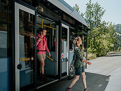 Zwei Frauen steigen aus einem Bus. Im Hintergrund sind grüne Berge zu sehen. 