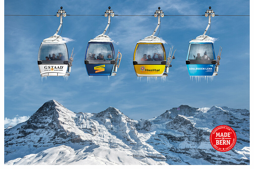 <p>Vier Gondeln (weiss, dunkelblau, gelb und königsblau) mit den Logos der Top4-Skiregionen Gstaad, Jungfrau Ski Region, Meiringen-Hasliberg und Adelboden-Len) hängen an einem Seil, darunter Bergpanorama.</p>