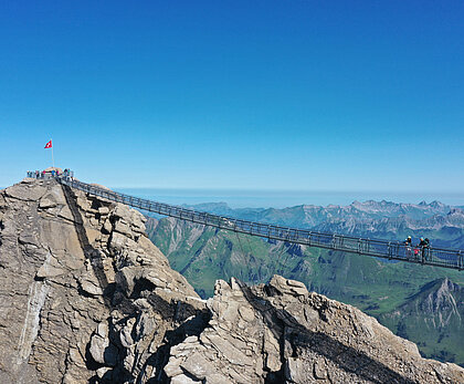 Eine Hängebrücke, welche zwei felsige Gipfel verbindet. Im Hintergrund ein sommerliches Bergpanorama.