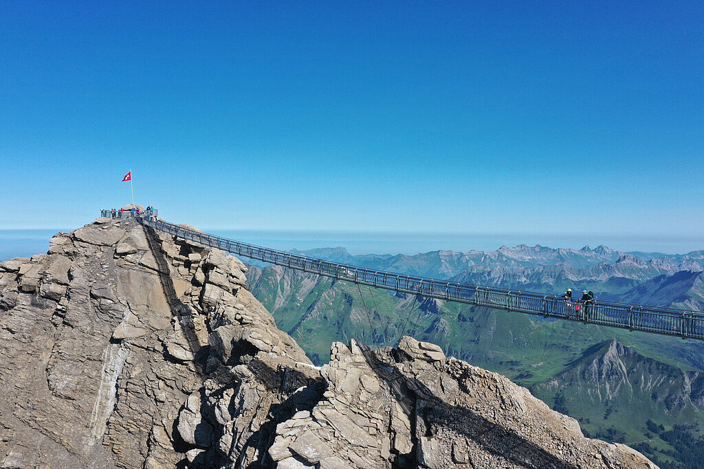 <p>Eine Hängebrücke, welche zwei felsige Gipfel verbindet. Im Hintergrund ein sommerliches Bergpanorama.</p>