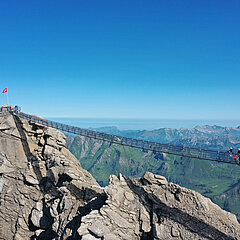 Un pont suspendu qui relie deux pics rocheux. En arrière-plan, un panorama de montagnes en été.