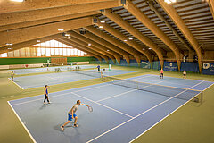 Eine Gruppe von Menschen spielen in einer grossen hölzernen Halle Tennis.