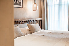 Bed with white blanket, white and beige pillows, a window with white and grey curtains at the top right, a lamp above the bed, a black and white picture on the wall.