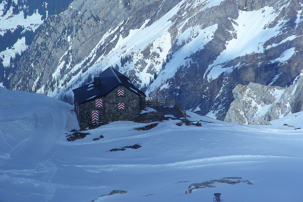 <p>Grau gemauerte Berghütte mit schwarzem Dach und geschlossenen rot-weiss gestreiften Fensterläden und einer Terasse in winterlicher Berglandschaft.</p>