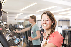 Eine junge Frau in lachsfarbenem T-Shirt steht in einem Fitness und lächelt in die Kamera.
